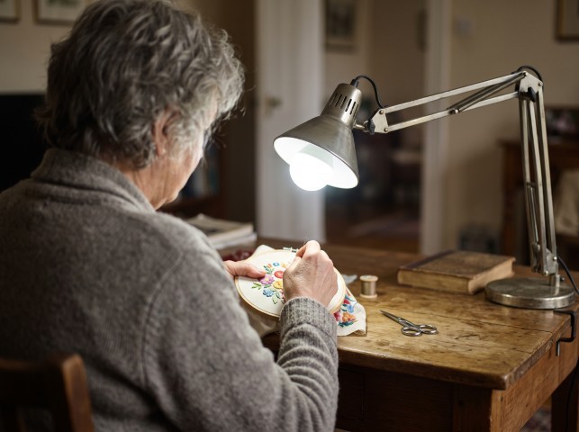 Older woman doing embroidery under a bright desk lamp at a wooden table.